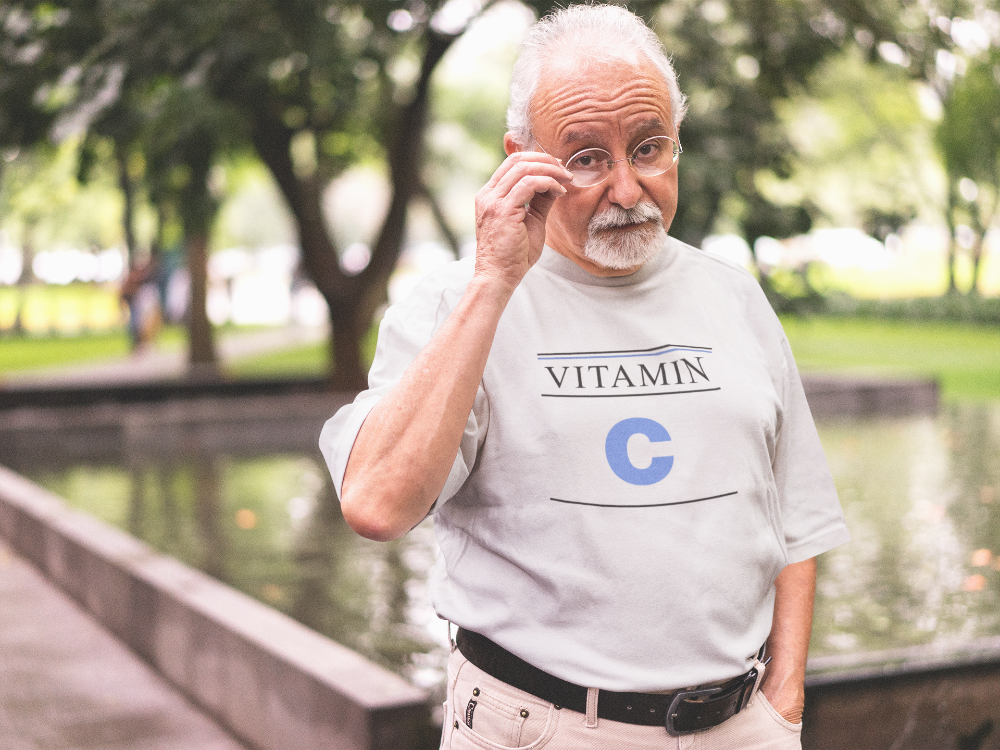 Man wearing a t-shirt with 'Vitamin C' printed on it, standing outdoors by a pond.